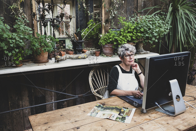 Senior woman wearing glasses, black top and white apron sitting at a wooden table, working on desktop computer.