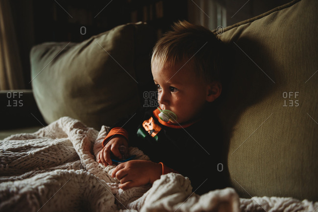 Toddler boy sitting on sofa with blanket and pacifiers