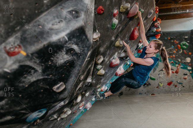 Side view of a woman hanging from an artificial climbing wall in an indoor bouldering gym. Female boulderer making her way up a bouldering wall.