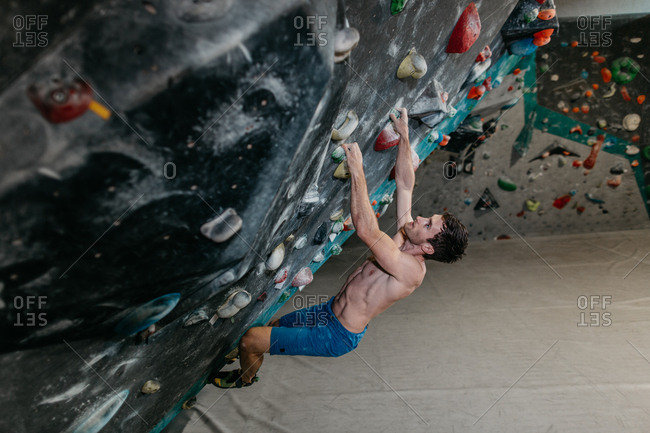 Side view of a man hanging from an artificial climbing wall in an indoor bouldering gym. Male boulderer making his way up a bouldering wall.