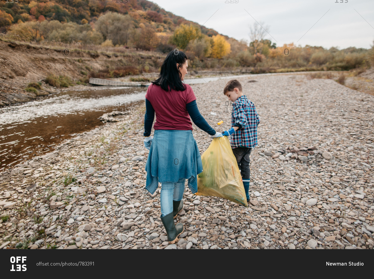 Back view of woman and her son walking along river beach collecting rubbish. Rear view of female volunteer and her child carrying plastic bag filled with garbage.