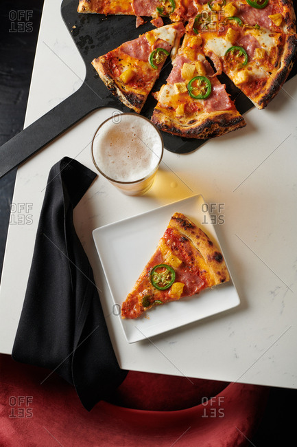 Slice of hawaiian pizza with pineapple and canadian bacon and sliced jalapeno on a square plate served with a pint of beer on a marble like table at a hip restaurant.