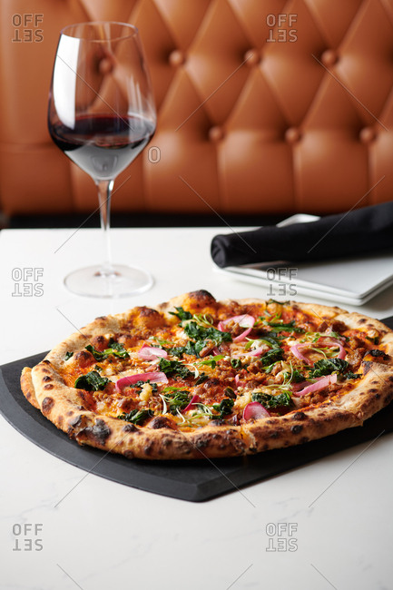 Sausage and Kale pizza with pickled red onions and micro greens made in a stone oven and served at a hip restaurant with a glass of red wine on a black pizza serving slab on a marble style table.