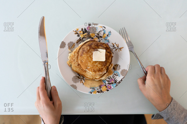 Person holding a knife and fork ready to eat a stack of pancakes topped with butter