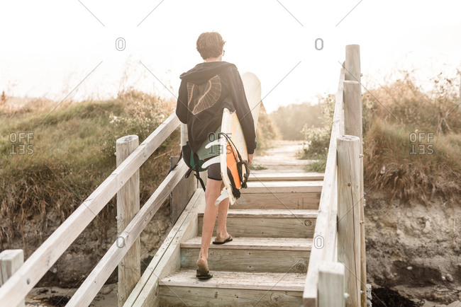 Rear view of teen walking up steps on beach with surfboard