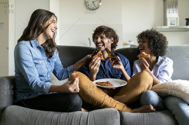 Cheerful friends sitting on couch drinking beer and eating pizza