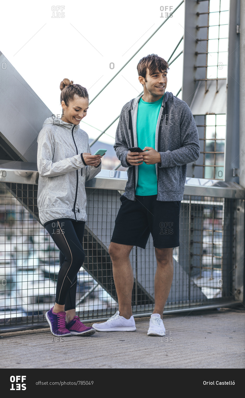 Young couple in athletic clothing standing on bridge using cell phones