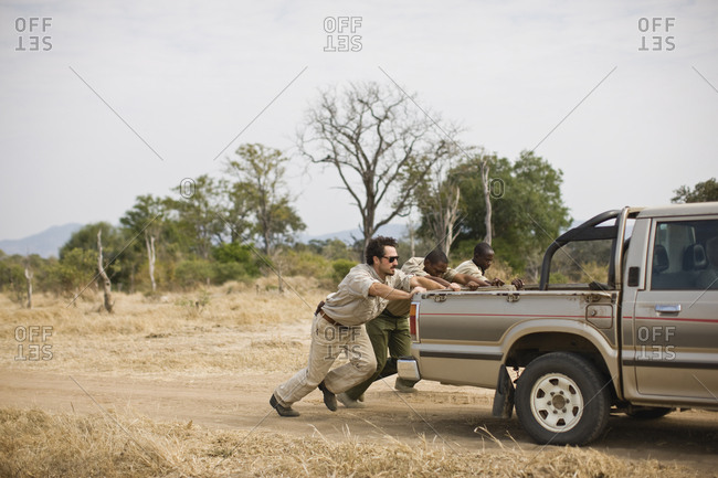 Three mid-adult men pushing a truck along a dirt road.