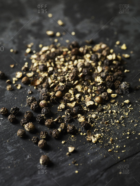 Whole black peppercorns on a piece of slate shot form an angle, with some crushed pepper scattered about.