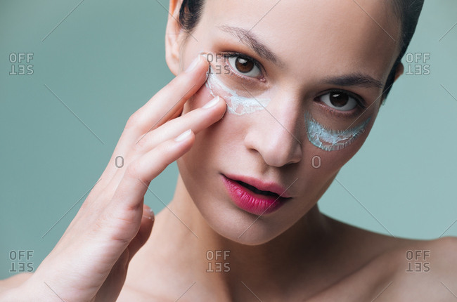 Close up of beautiful Caucasian woman putting cosmetic cream on her face and looking at camera.