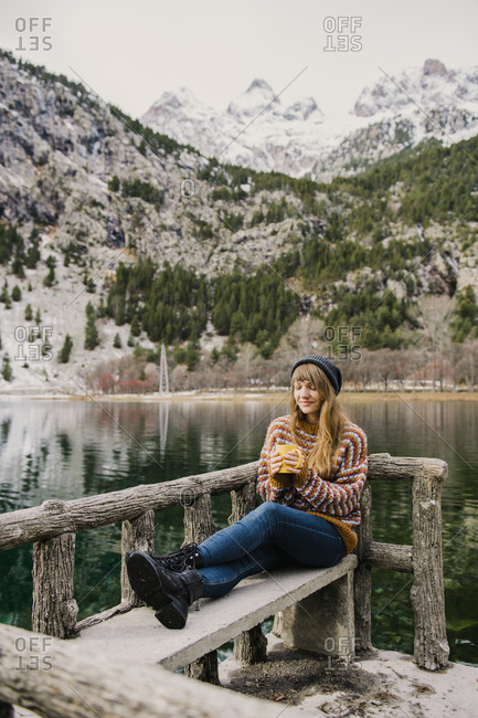 Young lady sitting on bench closed eyes near amazing view of water surface between high mountains with trees in snow in Pyrenees