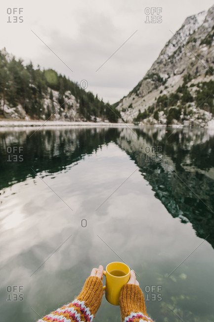 Crop hand of human holding yellow mug near amazing view of water surface between high mountains with trees in snow and cloudy heaven in Pyrenees
