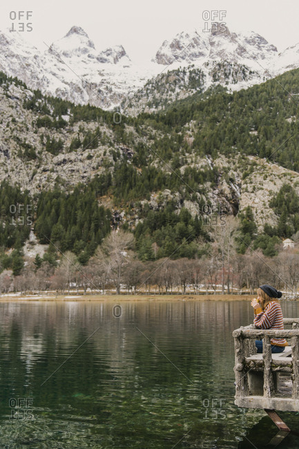 Side view of lady sitting on bench and looking at amazing view of water surface between high mountains with trees in snow and cloudy heaven in Pyrenees