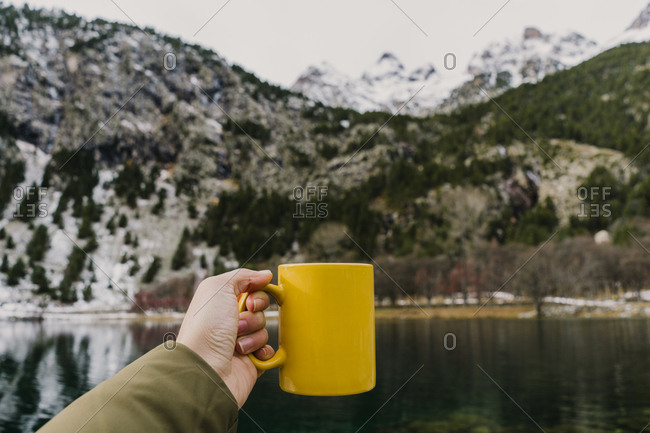 Crop hand of human holding yellow mug near amazing view of water surface between high mountains with trees in snow and cloudy heaven in Pyrenees