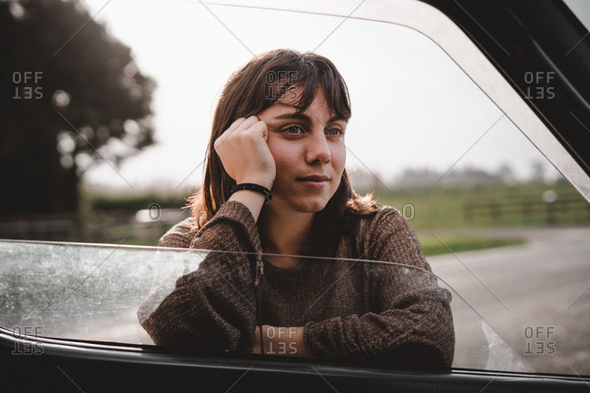 Dreamy young woman looking away and looking away while leaning on car door in Kaiate Falls, New Zealand