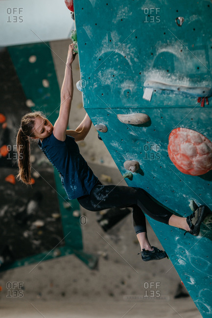 Female climber climbing up an artificial climbing wall in an indoor bouldering gym. Woman resting whilst making her way up a bouldering wall.