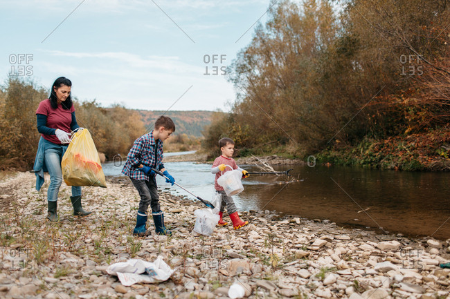Woman cleaning up plastic waste with her young sons on the beach. Female volunteer and her children picking up trash around river.