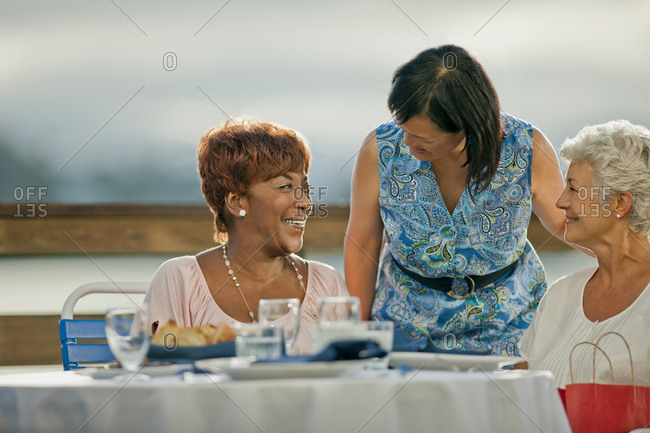 Two mature female friends out to lunch share a laugh with an acquaintance who has come to say hello.