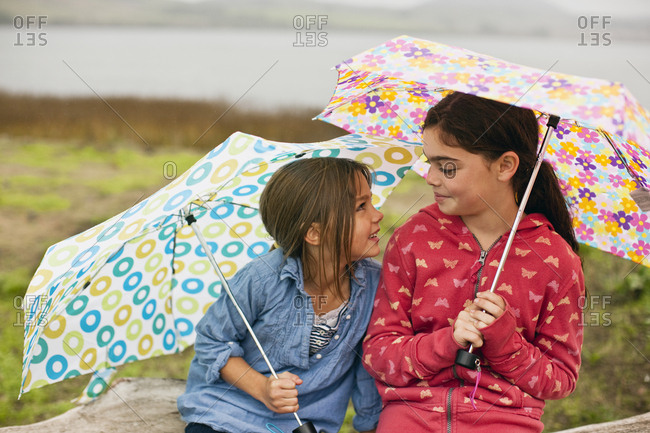 Two young girls smile and talk as they sit together on a fallen tree trunk while holding open umbrellas.