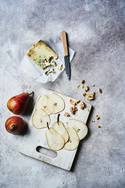 A view overhead of some pears, pear slices on a board with walnuts scattered on the board and falling off the surface. Some blue cheese is at the top of the shot crumbling apart with a knife to the right of it. Copy space is available around top, right and bottom of the image.