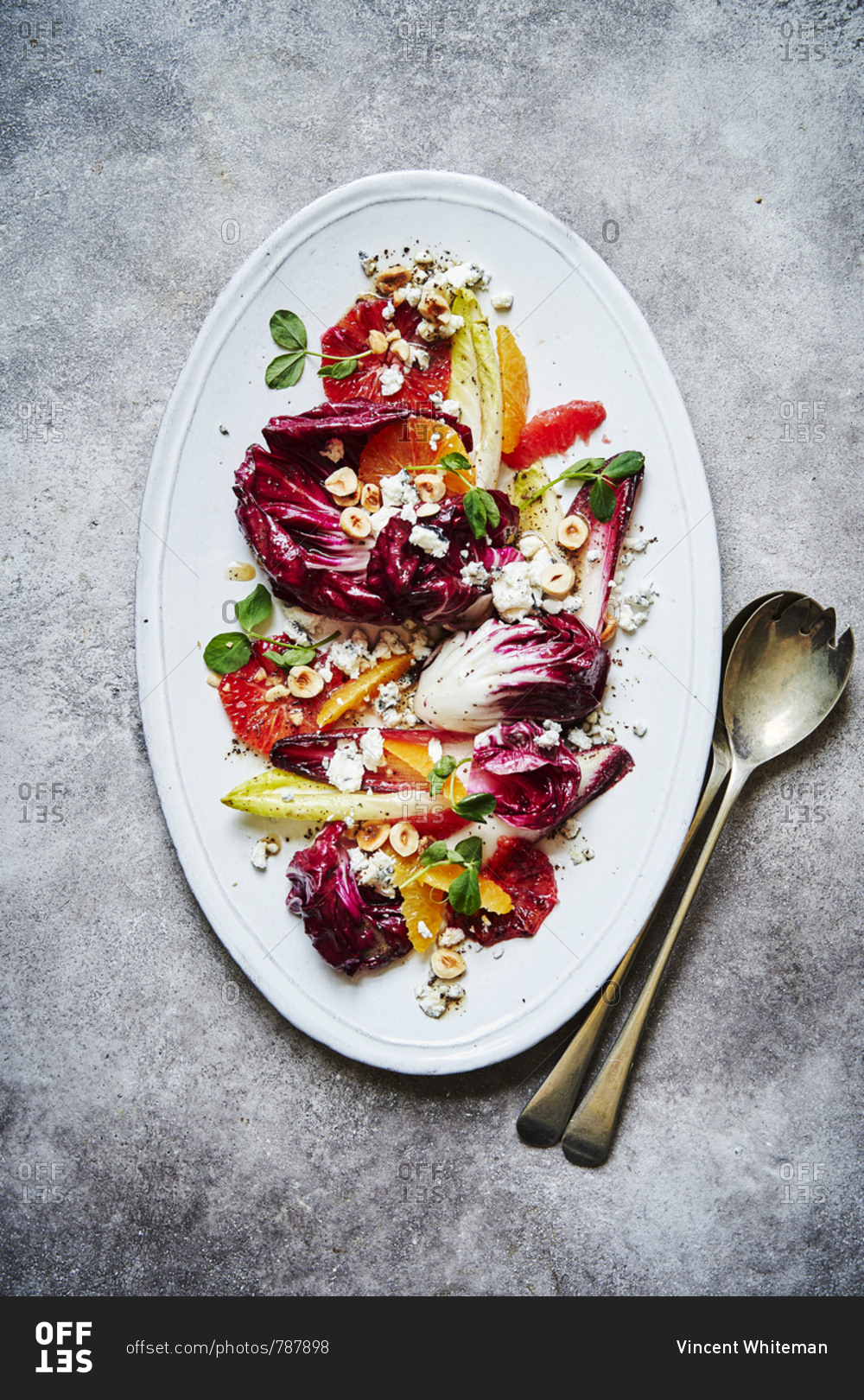 A citrus, chicory, hazelnut and goats cheese salad, shot from above served on a oval white dish with lots of punchy colours. Serving spoons are on the lower right of the image on top of a red cloth.