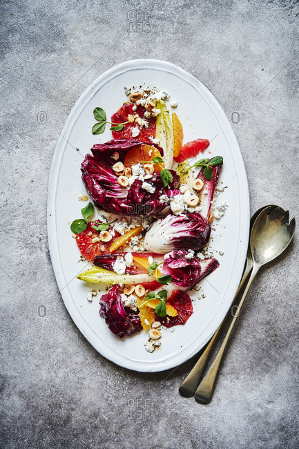 A citrus, chicory, hazelnut and goats cheese salad, shot from above served on a oval white dish with lots of punchy colours. Serving spoons are on the lower right of the image on top of a red cloth.