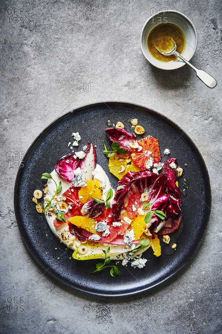 A citrus, chicory, hazelnut and goats cheese salad, sitting atop some yoghurt  and shot from above served on a dark round plate with lots of punchy colours. A ramekin of dressing with a spoon can be found a the top right.