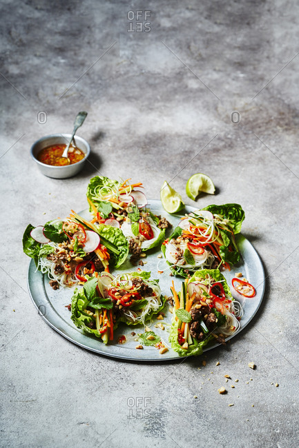Five Vietnamese lettuce wraps shot from mid three quarters with a wedge of lime and sauce at the top of the image. Ingredients include beef, rice noodles, carrot, cucumber, spring onions, chili, peanuts, coriander, mint and squeeze of lime.