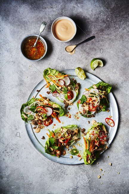 Five Vietnamese lettuce wraps shot from above with a wedge of lime and sauce and sriracha at the top of the image. Ingredients include beef, rice noodles, carrot, cucumber, spring onions, chili, peanuts, coriander, mint and squeeze of lime.
