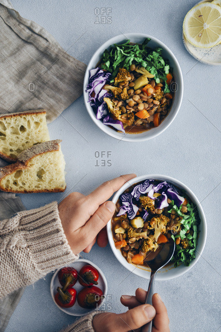 Vegan lentil stew with broccoli and carrot garnished with shredded red cabbage and chopped parsley served in three white bowls. Accompanied by bread slices, pickled chili and lemon slices on a grey background.