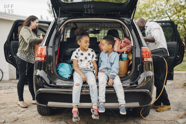 Full length of smiling siblings sitting on car trunk with parents standing in front yard