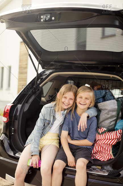Portrait of smiling siblings sitting in car trunk against house