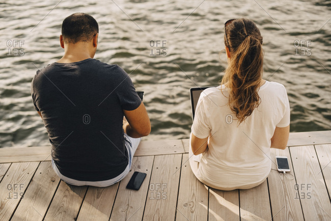 Rear view of couple using laptop while sitting on patio against lake