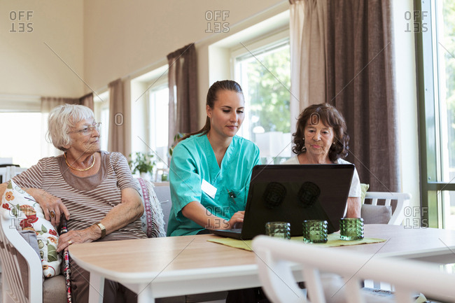Young nurse using laptop while sitting amidst senior women at nursing home