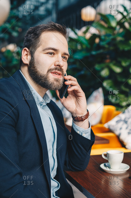 Attractive and urban young man sitting in a garden cafe bar, having a friendly conversation over his mobile phone during a break from work. Vertical image.