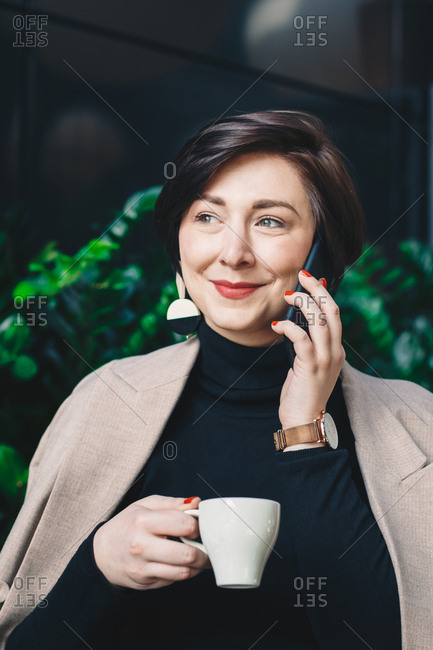 Portrait of a young fashion blogger sitting in a garden bar during a business break, drinking coffee and talking on her smartphone. Vertical image.