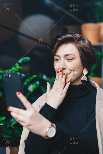 Portrait of attractive and urban young social media influencer sitting in a garden bar full of plants during business downtime, live streaming with online followers over her phone. Vertical image.