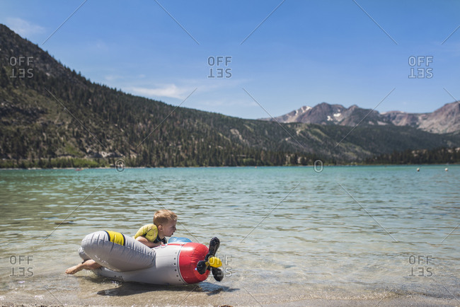 Cute playful boy lying on inflatable airplane by lake against mountains during summer