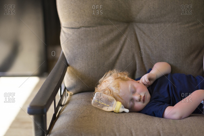 Cute Baby Boy Sleeping By Milk Bottle On Couch At Home Stock Photo