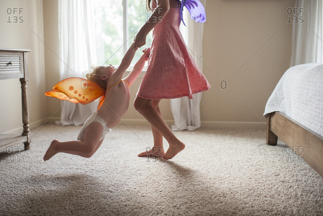 Low section of playful sister with costume wings holding brother's hands while spinning him in bedroom at home