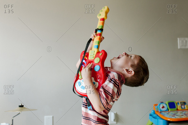Cute happy boy singing while playing toy guitar against wall at home
