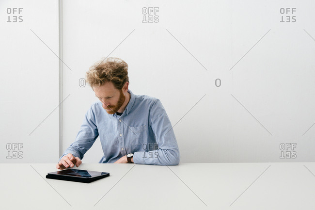 Thirty something bearded man looking thoughtfully on his tablet sitting at an empty light colored desk.