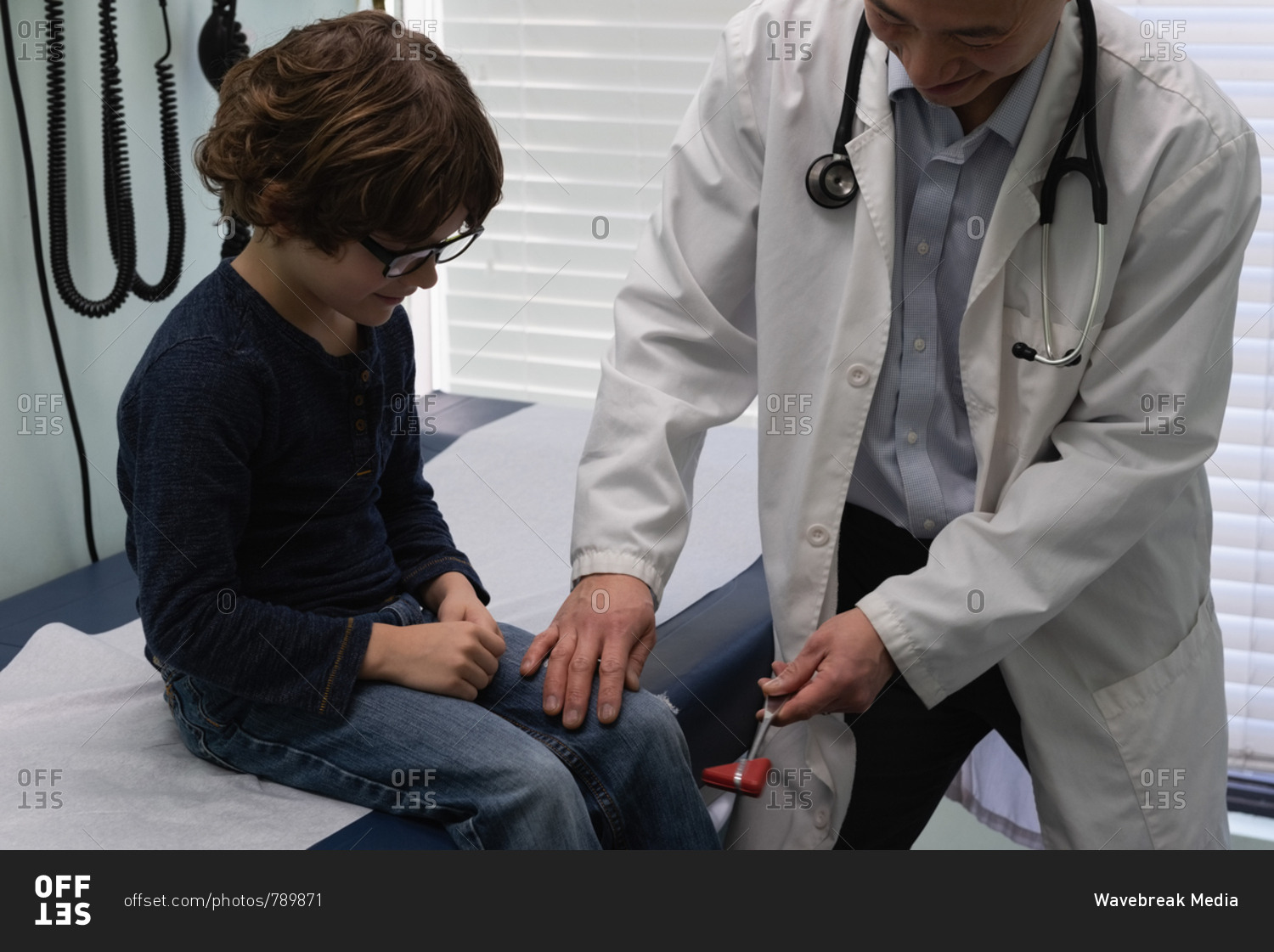 Front view of young asian male doctor examining caucasian boy patient knee in a clinic. Male doctor using reflex hammer