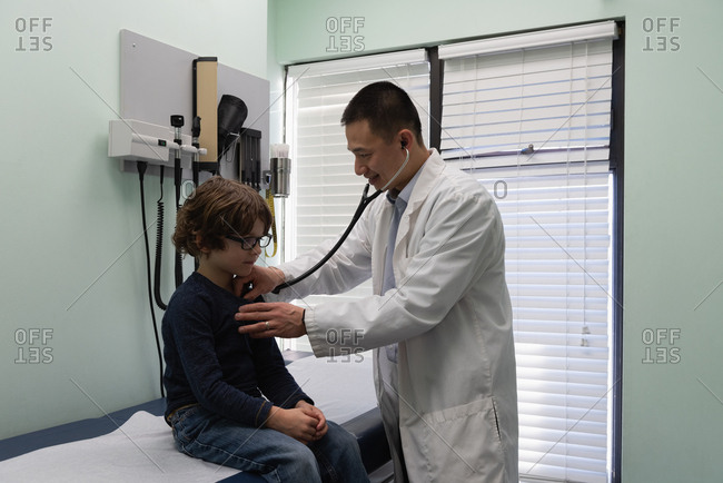 Side view of young asian male doctor examining caucasian boy patient chest in a clinic. Male doctor using stethoscope
