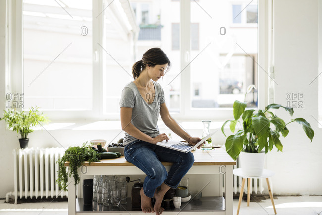 Woman sitting on kitchen table- searching for healthy recipes- using laptop