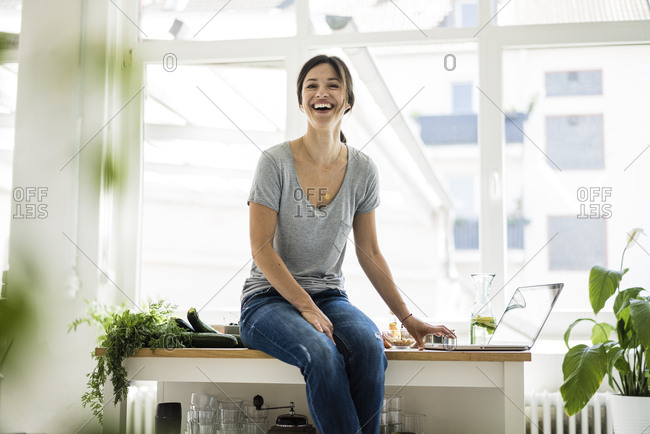 Woman sitting on kitchen table- searching for healthy recipes- using laptop
