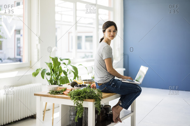 Woman sitting on kitchen table- searching for healthy recipes- using laptop