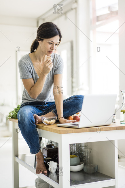 Woman sitting on kitchen table- searching for healthy recipes- using laptop