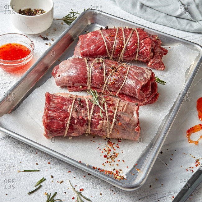 Homemade natural raw beef roulades on a baking tray with spices and red sauce on a white wooden background.
