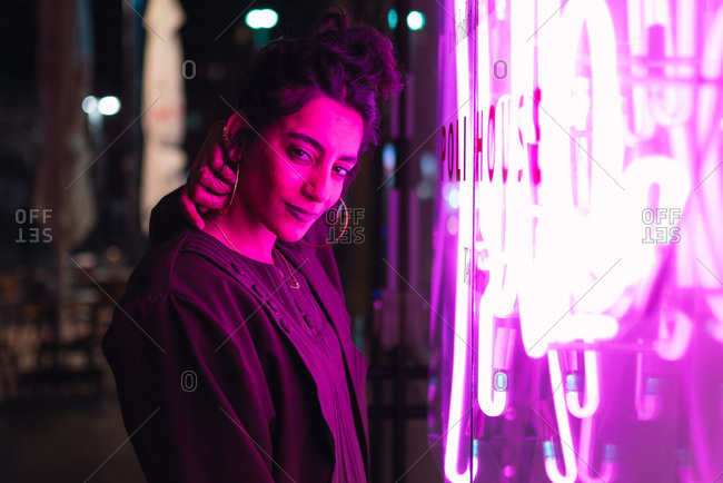 Beautiful trendy young hipster with earrings looking at camera near pink illumination on street in Tel Aviv, Israel
